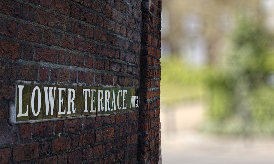 A stone pathway leading through well-maintained flowerbeds filled with pink and white blossoms, extending towards a historic brick townhouse with large sash windows and a central entrance. The buildin