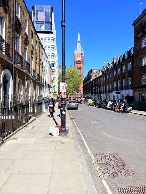 A city street scene with a wide concrete sidewalk on the left, lined with a black metal railing and a few pedestrians carrying shopping bags. The sidewalk is clean and free of debris. On the right sid