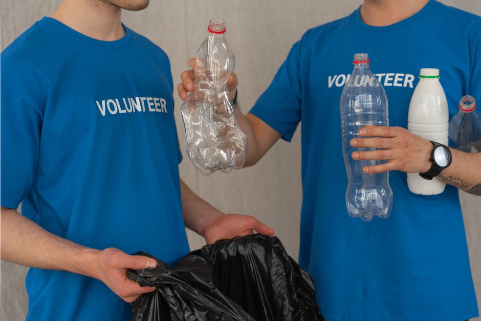 A young woman with curly hair and a light brown complexion, dressed in a blue volunteer t-shirt, is standing next to a transparent recycling bin marked with green and orange recycling symbols for pape