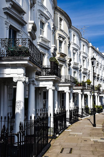 Image of a row of white Victorian-style terraced houses in Hampstead, featuring ornate architectural details, black wrought-iron balconies, and black iron fences along the pavement. The buildings have
